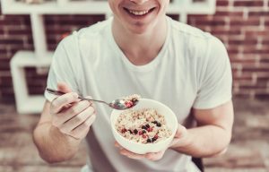 Handsome young guy is smiling while eating porridge with berries in kitchen at home