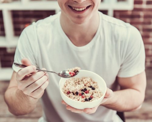 Handsome young guy is smiling while eating porridge with berries in kitchen at home