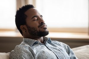 Tranquil calm young african american man resting with eyes closed taking deep breath of fresh air enjoy comfort relaxing on couch feel peace mind and stress relief meditate at quiet home sit on sofa