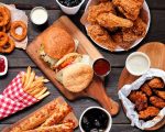 Table scene of assorted take out or delivery foods. Hamburgers, pizza, fried chicken and sides. Top down view on a dark wood banner background.