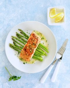 Fresh grilled wild coho salmon filet with lemon butter sauce on a bed of steamed organic asparagus. White dishes on pale blue background. Healthy eating concept.