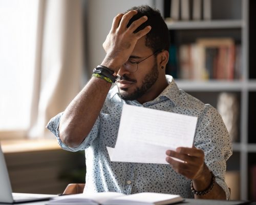 Stressed african businessman sit at desk hold paper letter reading bad awful negative news got fired from work. Dismissal notice, financial problems debt notification from bank, unpaid invoice concept