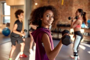 African american girl smiling at camera while exercising using dumbbell in gym together with female trainer and other kids. Sport, physical education concept. Horizontal shot. Selective focus
