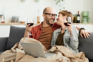 Happy mature couple sitting on sofa with glasses of red wine using laptop computer and laughing