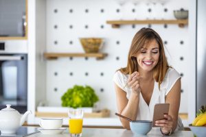 Beautiful young woman having breakfast in the kitchen. Beautiful cute smiling woman is using smart phone on the kitchen at home. Woman Eating Breakfast Whilst Using Mobile Phone