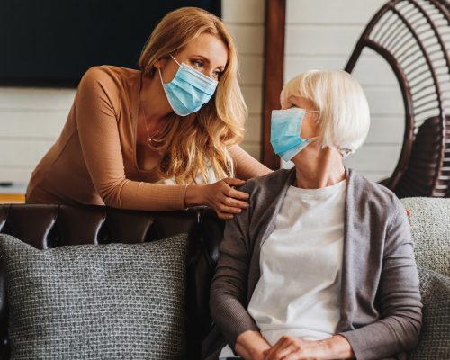 Senior woman in medical mask with social worker visiting her at home