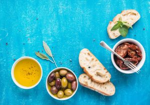 Mediterranean snacks set. Olives, oil, sun-dried tomatoes, herbs and sliced ciabatta bread on over blue painted background, top view, copy space