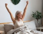 Active happy mature female wake up from good healthy sleep stretching sitting in bed at home, smiling positive senior woman awaken in comfortable bedroom feel optimistic welcome new day