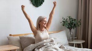 Active happy mature female wake up from good healthy sleep stretching sitting in bed at home, smiling positive senior woman awaken in comfortable bedroom feel optimistic welcome new day