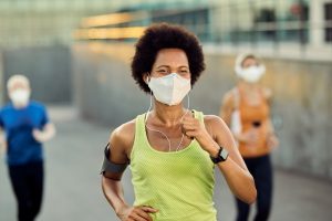 Happy African American female runner wearing protective face mask while jogging outdoors during coronavirus epidemic. There are people in the background.