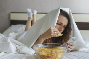 Girl eating chips in bed and watching the news in phone. Lying on a white bed under a blanket