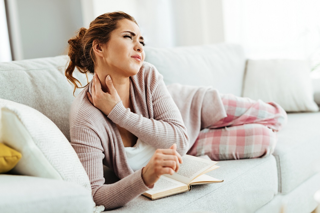 Young woman having neckache while lying down on sofa and reading book in the living room.