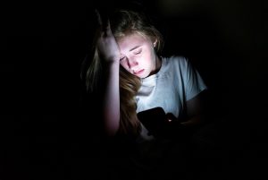 The image displays an upset girl sitting in the dark while using her smartphone. The light from the screen is illuminating her face.
