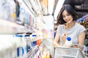 Young Asian beautiful woman holding grocery basket walking in supermarket. She is choosing daily milk product picking up from shelf. Seen from side while she looking at products. Shopping concept.