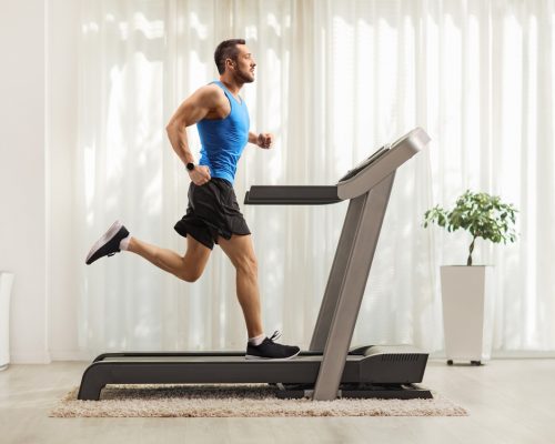 Full length profile shot of a young man running on a treadmill at home