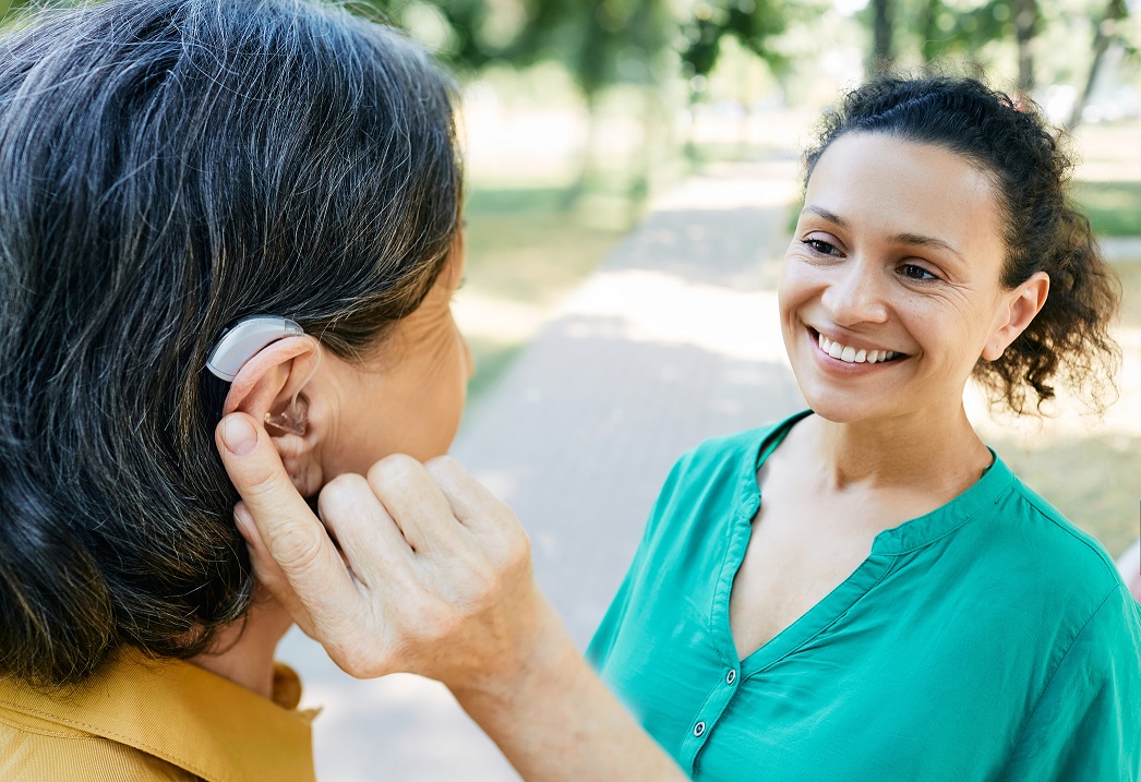 Mature woman with a hearing impairment uses a hearing aid to communicate with her female friend outdoor. Hearing solutions