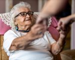 Female home carer supporting old woman to stand up from the armchair at care home