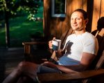 Man happy sitting relaxing on rocking chair lounge on porch of house in morning wooden cabin cottage drinking coffee or tea from cup mug