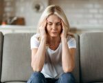 Middle aged blonde woman sits on couch at living room holding her head with her hands, feels unhappy because of headache, personal troubles, illness or bad news, she need psychological or medical support