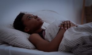 Insomnia, sleep problems, health care and bed sleep. Depressed young african american young woman lies on bed with blanket with open eyes, in dark bedroom interior, copy space, profile, panorama