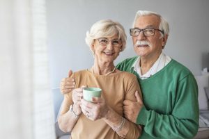 Enjoying time together. Side on waist up portrait of happy senior woman and man staying embracing near window at home interior. Portrait of senior couple standing near window (Enjoying time together. Side on waist up portrait of happy senior woman and
