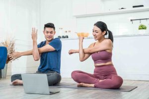 Young Asian sporty couple watching live or video tutorial practice yoga lesson at home together. healthy man and woman Stretching and balancing body on exercise mat. wellness relaxing lifestyle
