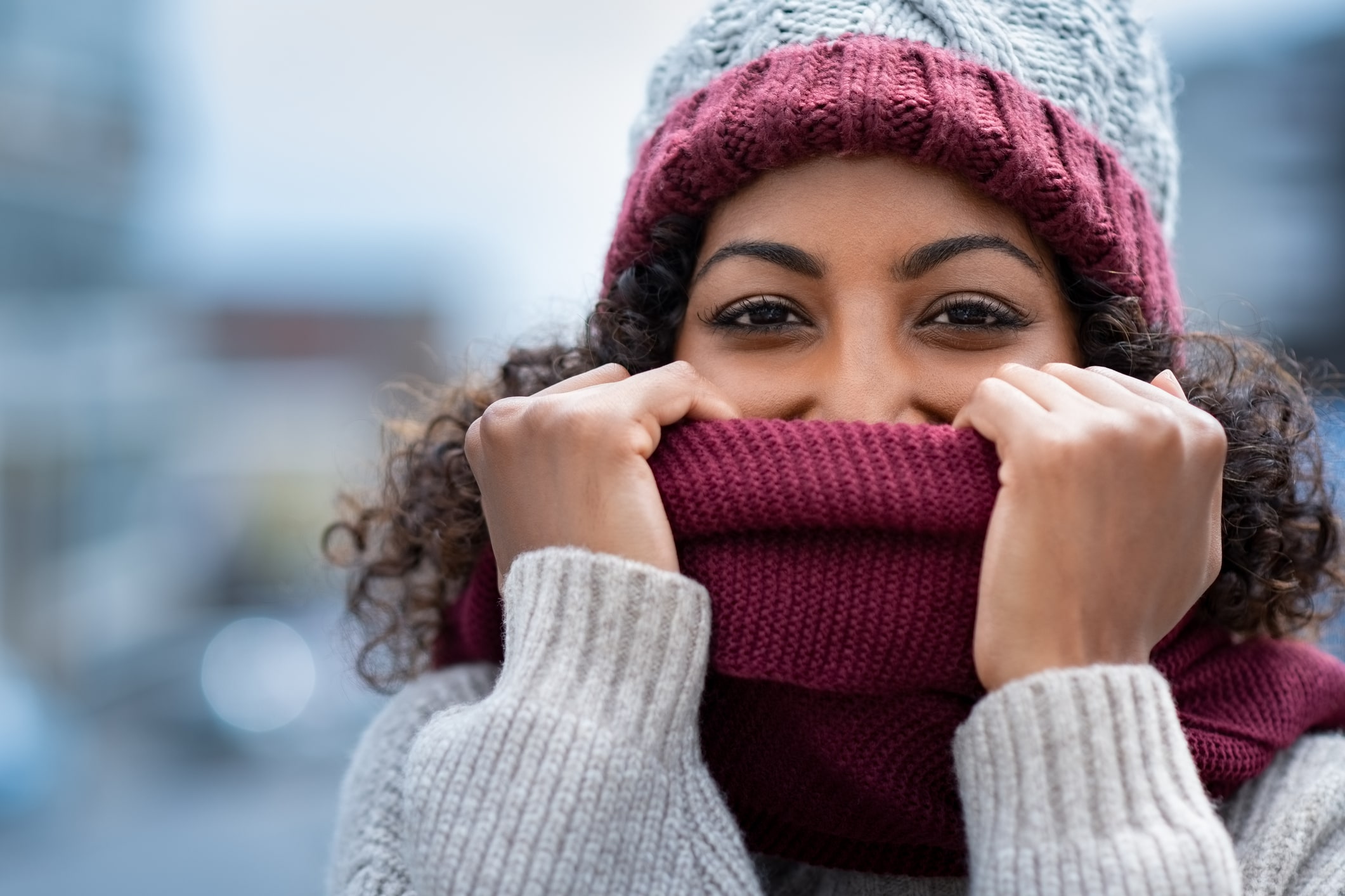 Closeup face of happy african girl holding woolen scarf with hands over nose to protect from the frost. Portrait of beautiful young black woman in warm winter knitted clothes covering her face and looking at camera.
