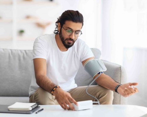 Young arab man sitting on couch, checking blood pressure and taking notes at home. Indian guy in white t-shirt using modern tonometer, experiencing hypertension caused by stress, copy space (Young arab man sitting on couch, checking blood pressure and