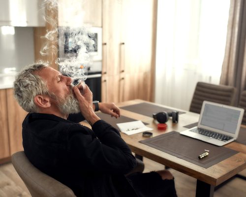 Middle-aged man smoking marijuana cigarette or joint, sitting in the kitchen and writing song using laptop. Marijuana grinder, lighter, headphones and weed on the table. Cannabis legalization concept