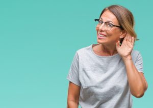 Middle age senior hispanic woman wearing glasses over isolated background smiling with hand over ear listening an hearing to rumor or gossip. Deafness concept.