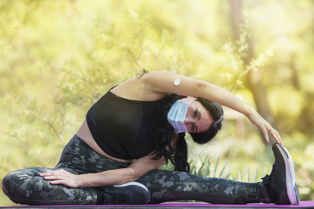 Adult woman with sportswear and a glucose sensor device on her arm doing stretching on a purple mat in the park