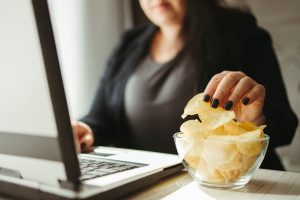 Compulsive overeating, mindless snacking, junk food, unhealthy meals. Woman eating chips from bowl at her workplace