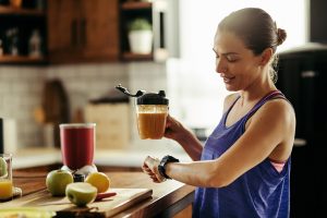 Smiling athletic woman drinking healthy smoothie and checking the time on her wristwatch in the kitchen.
