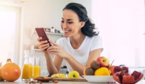 Smiling cute woman is using smartphone in the kitchen at home while cooking fruits vegan salad