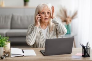 Portrait Of Shocked Mature Woman Talking On Smartphone Grabbing Head In Amazement And Surprise, Free Copy Space. Surprised Concerned Senior Lady Hearing Unexpected News, Sitting At Desk With Laptop