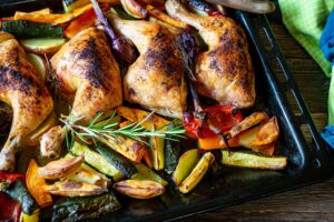 Homemade fresh cooked healthy family meal with roasted chicken legs and oven baked vegetables served on a baking tray on wooden table. Closeup view
