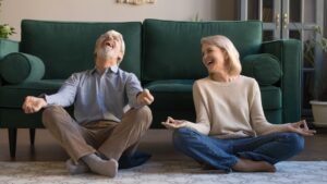 Laughing funny elderly spouses sitting in living room on floor in lotus position practice meditation distracted from yoga exercise joking feels overjoyed, healthy active lifestyle of retirees concept