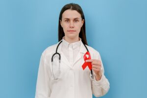 Female doctor holds red ribbon, looking at camera, wears uniform and stethoscope, isolated on blue wall in studio. Blood transfusion and donation. Hemophilia, health concept. World AIDS and HIV day