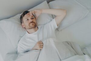 Young upset and tired bearded man lying on bed with open eyes and cannot sleep, male having insomnia problem or sleeping troubles. Bedtime and rest difficulty concept