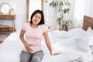 Menstrual Cramps. Chinese Girl Having Pelvic Lower Abdominal Pain Having Reproductive Health Problem Sitting In Bed At Home. Copy Space