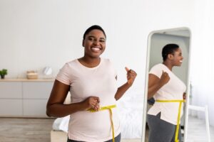 Plus size black woman measuring waist with tape, showing results of slimming diet, standing in front of mirror, gesturing thumb up at home. Young lady promoting healthy nutrition for weight loss