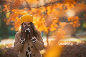 Hello autumn. modern 40 years old woman in brown coat and yellow hat with napkin blowing nose outdoors in the city park in autumn.