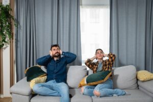Young couple is sitting on a sofa in their apartment looking up and holding their hands to plug their ears as a neighbor upstairs is having a party and playing loud music or renovating the apartment