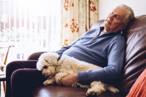 Senior man asleep at home on his sofa with his pet dog curled up next to him.