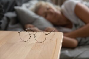 Close up of round glasses lying on bedside table in bedroom, senior woman sleep in bed in background, mature grandmother take off spectacles taking nap, elderly eyesight problem correction concept