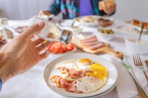 Sprinkles eggs and bacon with salt from a shaker. An excess of sodium chloride mineral can lead to the development of heart and metabolism diseases