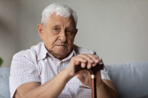 Head shot portrait of upset old senior hoary male pensioner with walking disability holding hands on wooden cane, sitting alone on couch at home, suffering from physical disease, ageing concept.