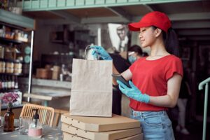 Side view of young attractive woman in latex gloves packing food in paper bags for delivery through city