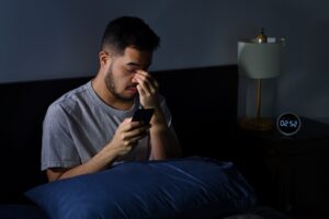 Young asian man tired with sore eyes from using smart phone while lying in bed at night.Young man has insomnia and can't sleep