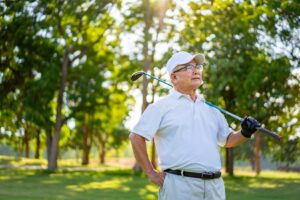 Portrait of Smiling Asian senior man golfer holding golf club standing on golf course in summer sunny day. Healthy elderly male enjoy outdoor lifestyle activity sport golfing at golf country club.